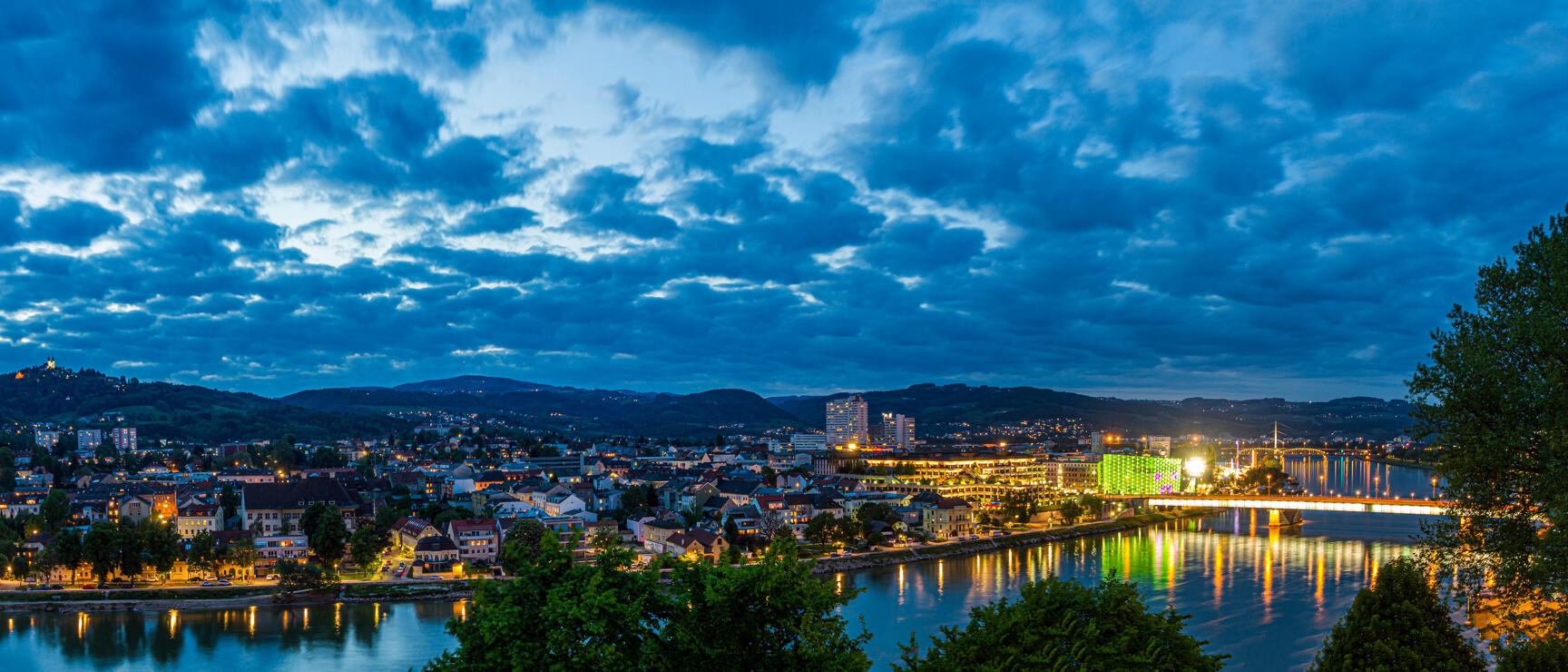 Panoramic view of Linz at night, Danube with bridge and illuminated Ars Electronica Center, cloudy evening sky.