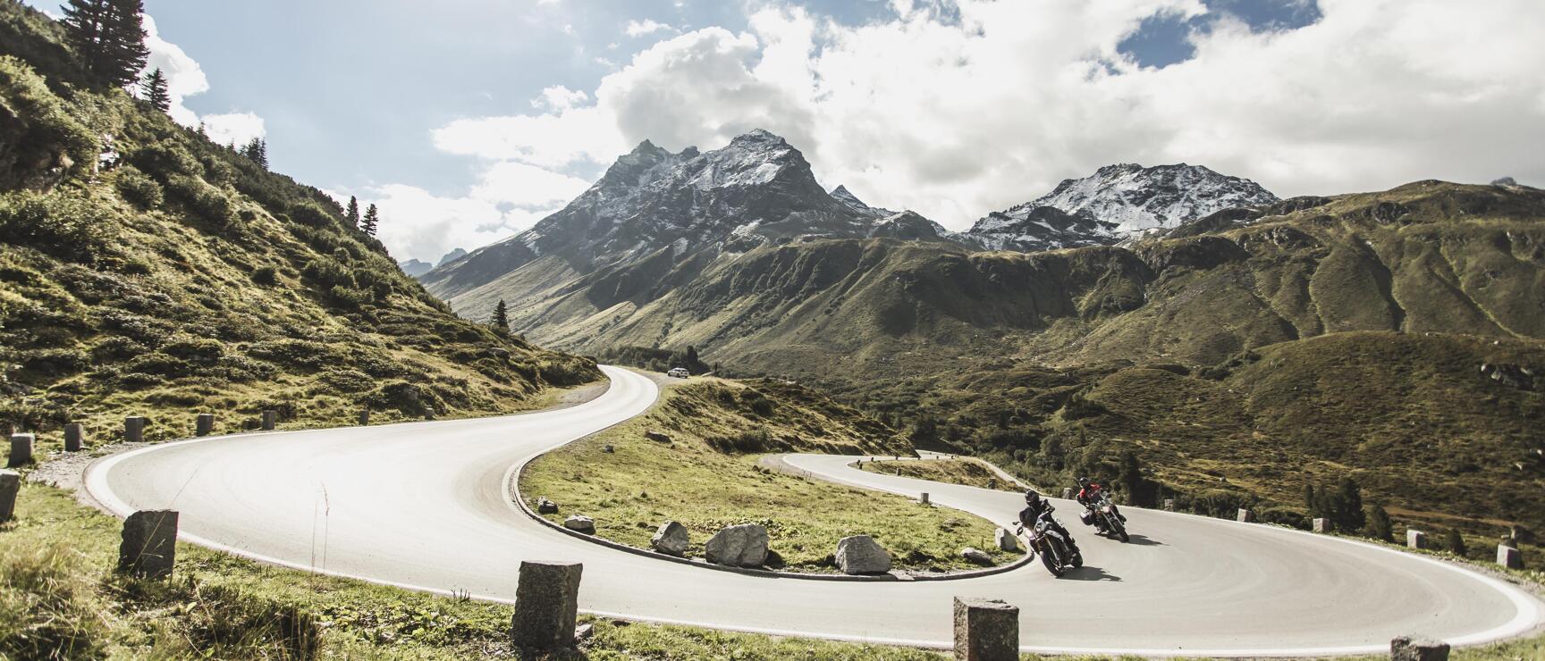 Zwei Motorräder auf einer Serpentinenstraße in den Alpen, schneebedeckte Gipfel im Hintergrund.