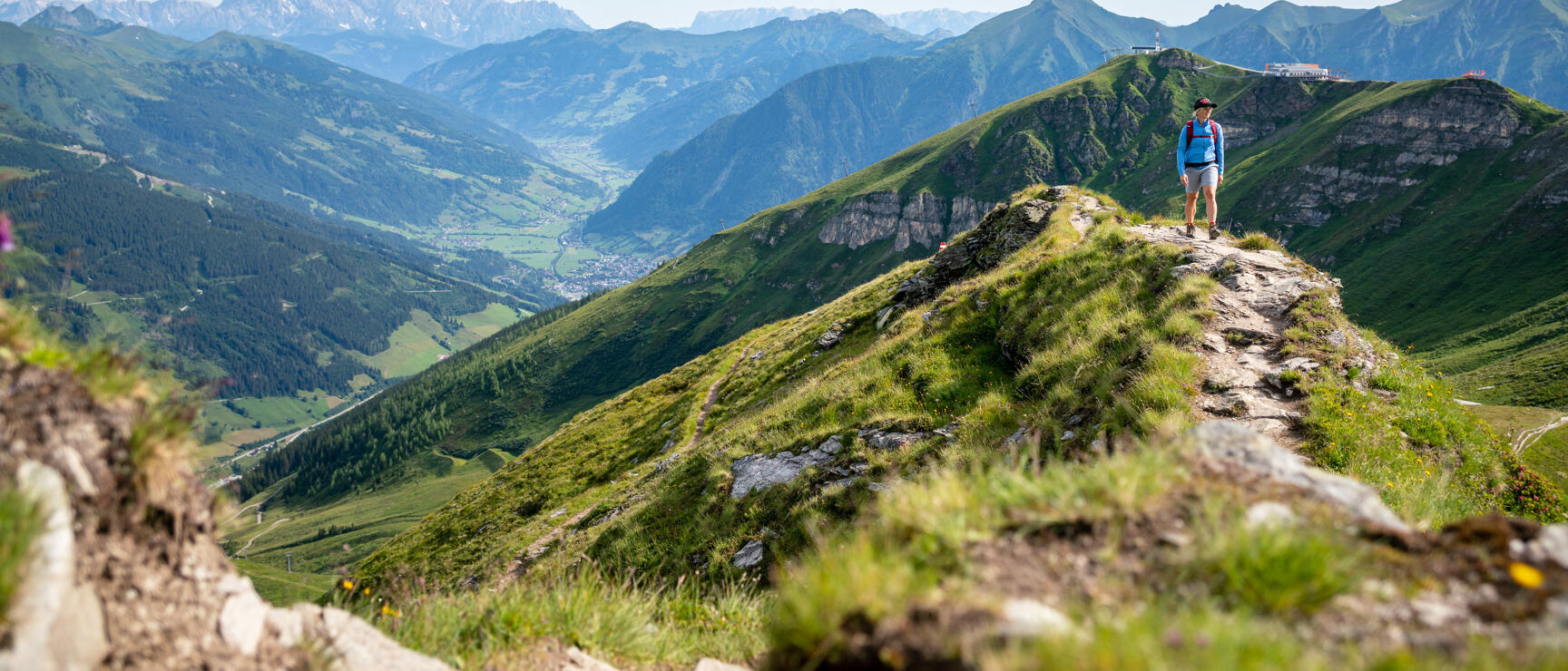 Hiker on narrow ridge with green mountain peaks and valley settlement in background.