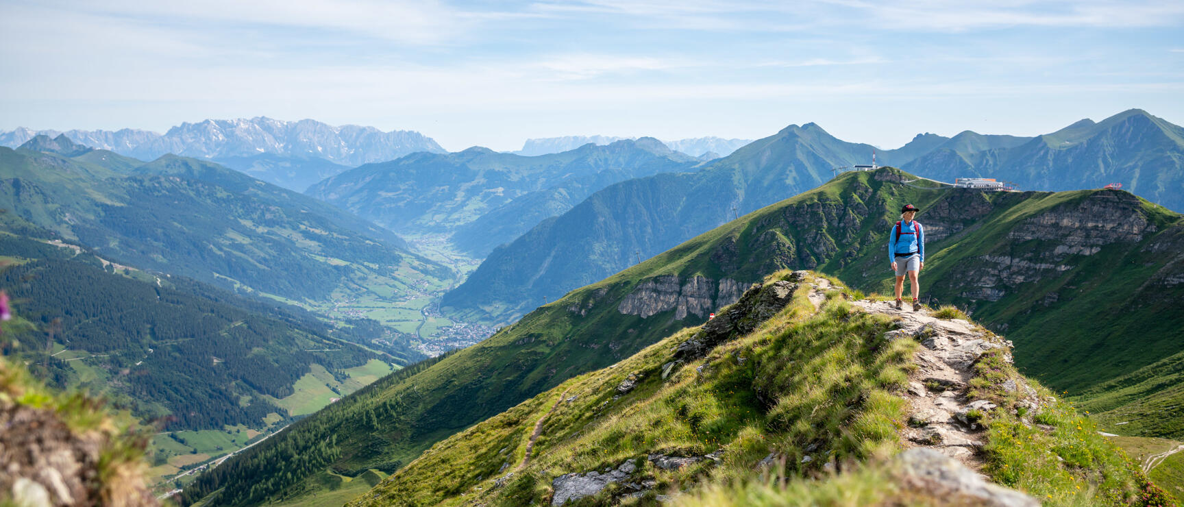 Hiker on narrow ridge with green mountain peaks and valley settlement in background.