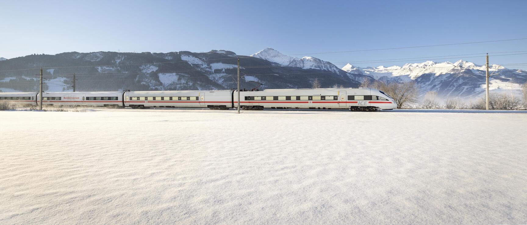 ICE-Zug fährt durch verschneite Winterlandschaft, schneebedeckte Alpen im Hintergrund, blauer Himmel.