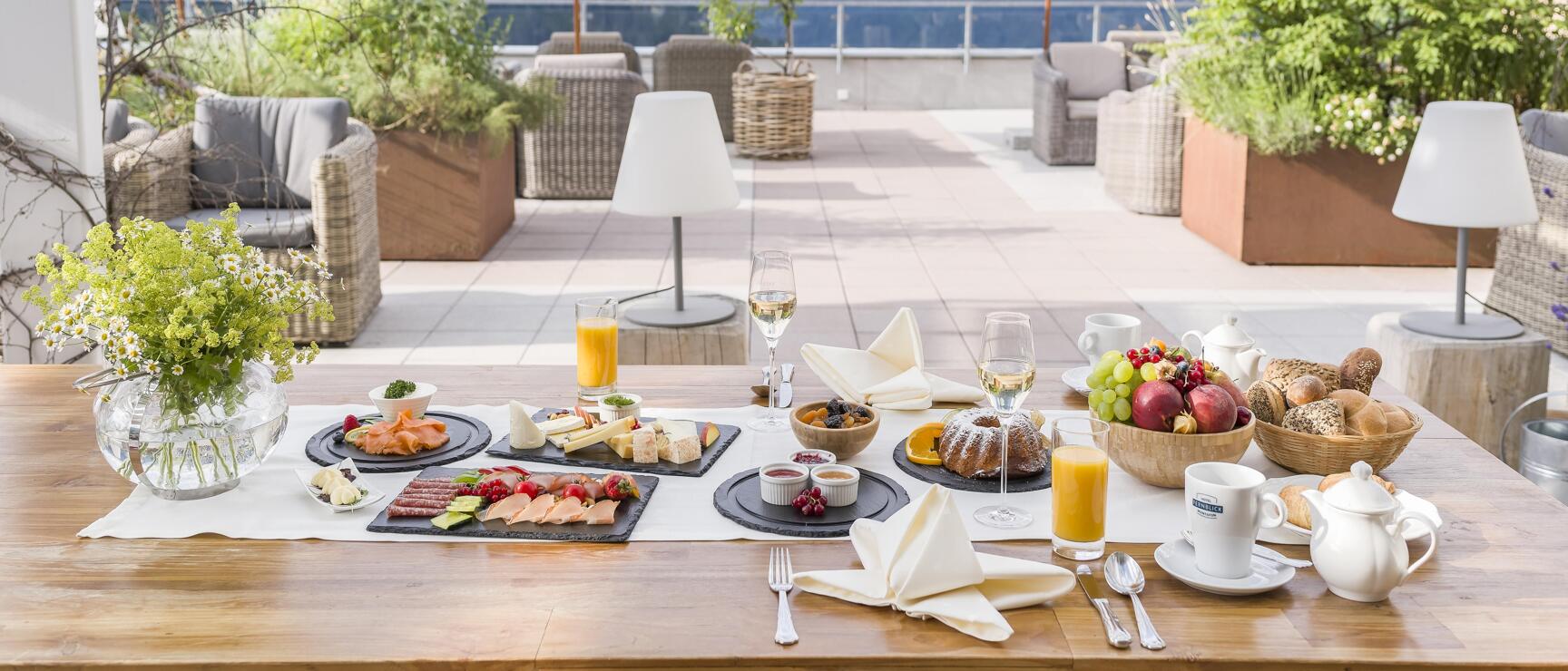 Breakfast table set on hotel terrace with cheese, cold cuts, fruit, juice and flowers, Alpine panorama in the background.