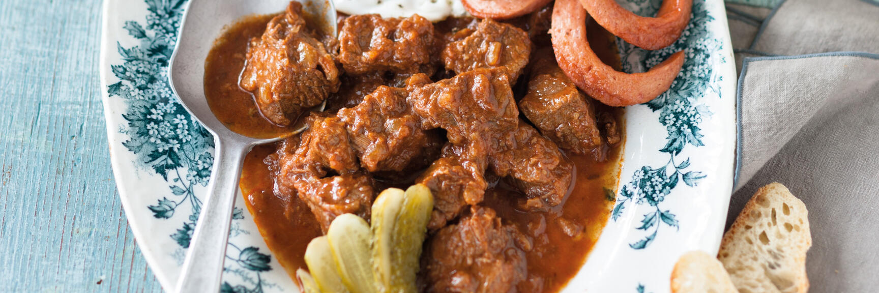 Goulash with fried egg, sausages and pickled cucumbers on plate, with bread and beer mug beside.