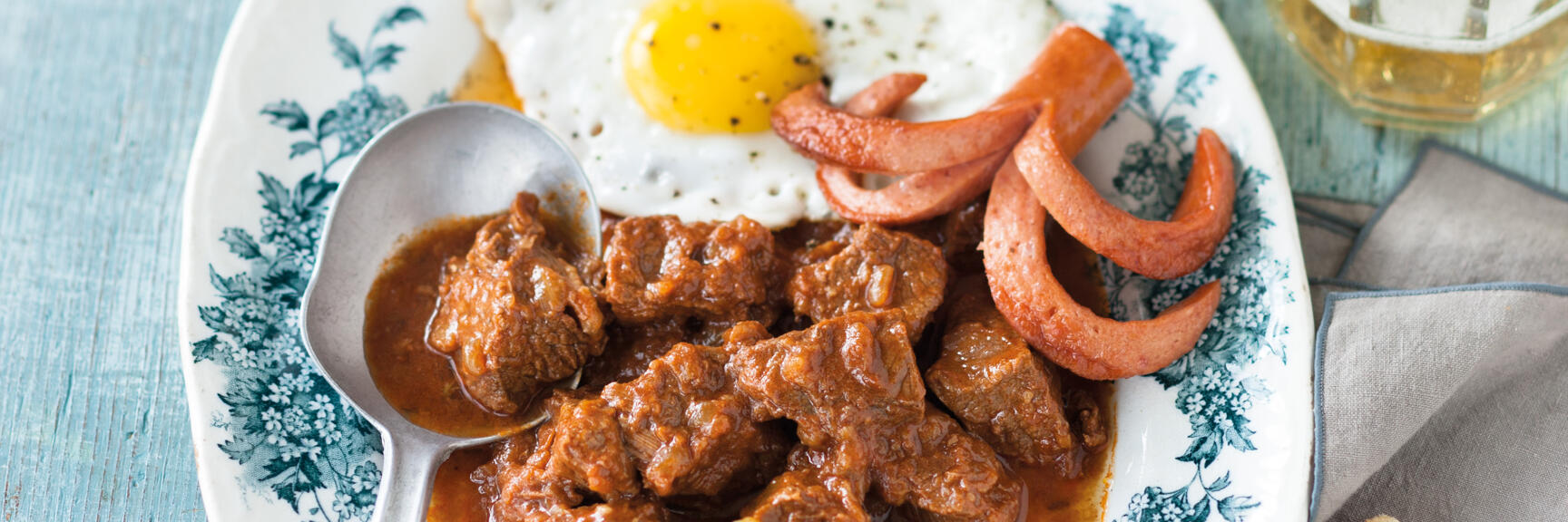 Goulash with fried egg, sausages and pickled cucumbers on plate, with bread and beer mug beside.