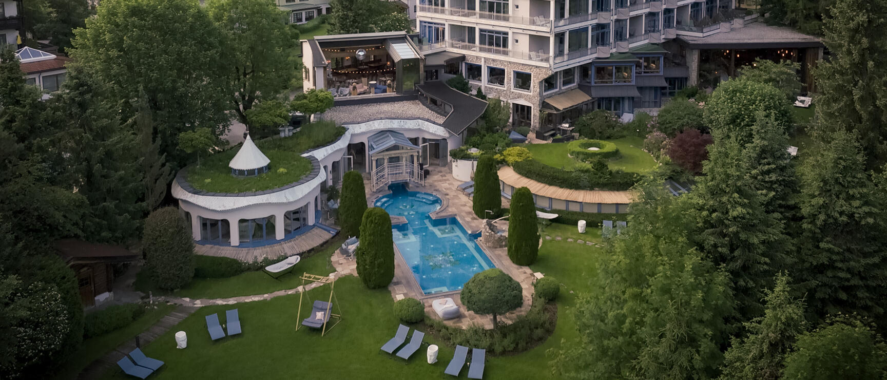 Aerial view of a hotel with outdoor pool, garden landscaping and sun loungers, surrounded by alpine village and green mountains.