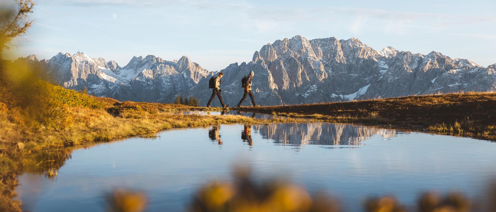 Two hikers with backpacks at mountain lake, snow-covered rocky peaks and water reflection.