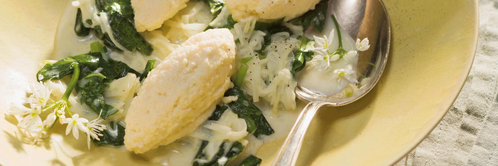 Nockerln mit Spinat und weißen Blüten in cremiger Sauce, serviert in gelbem Teller mit Löffel.