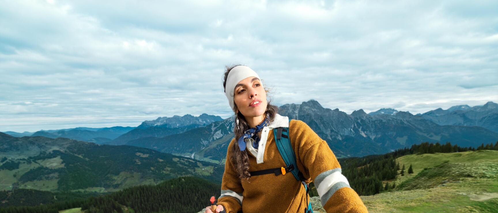 Woman with trekking poles and backpack on a rocky outcrop, vast mountain landscape in the background.