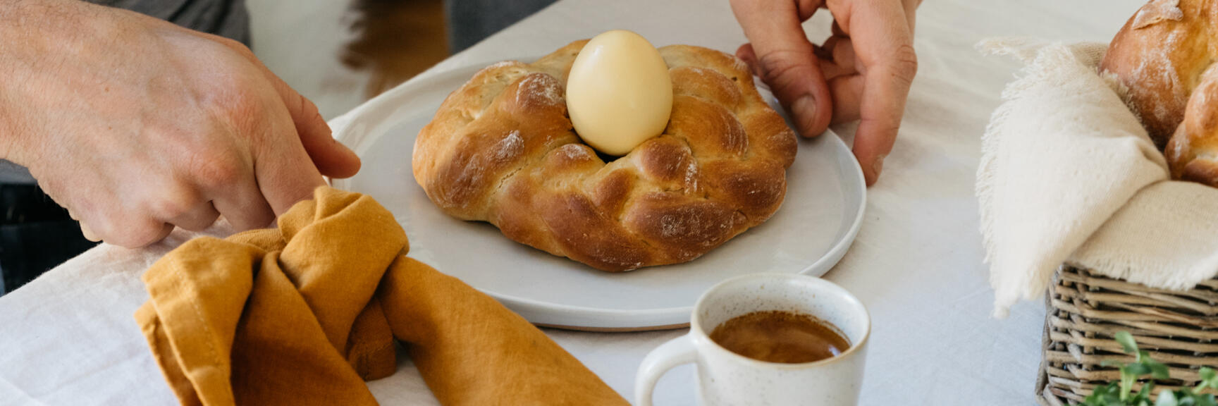 Table set with two Easter breads with egg, coffee, jam and cress, two hands offering bread.