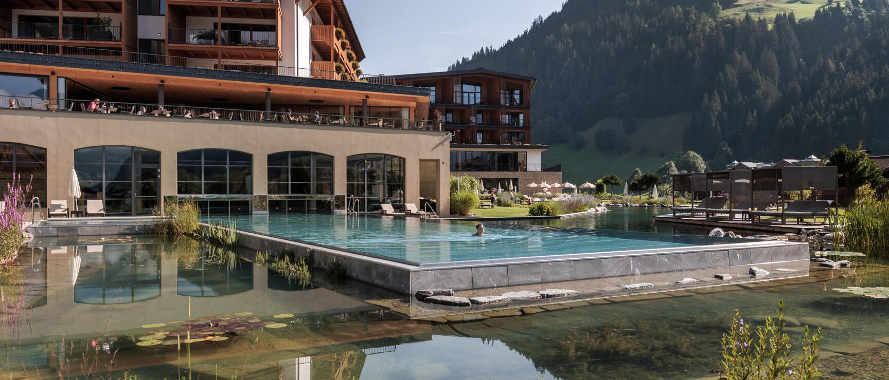 Outdoor pool of an alpine hotel with natural pond, sunbathing area and forested mountains in the background.