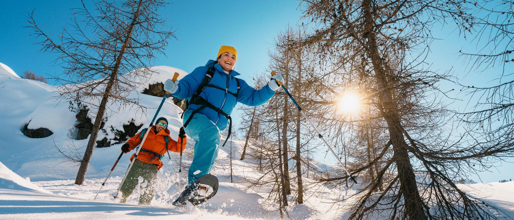 Zwei Personen beim Schneeschuhwandern durch eine sonnige, verschneite Winterlandschaft mit locker stehenden Bäumen. Die klare Bergluft, glitzernder Schnee und die dynamische Bewegung vermitteln Leichtigkeit, Naturerlebnis und Freude am Winter in Österreich.