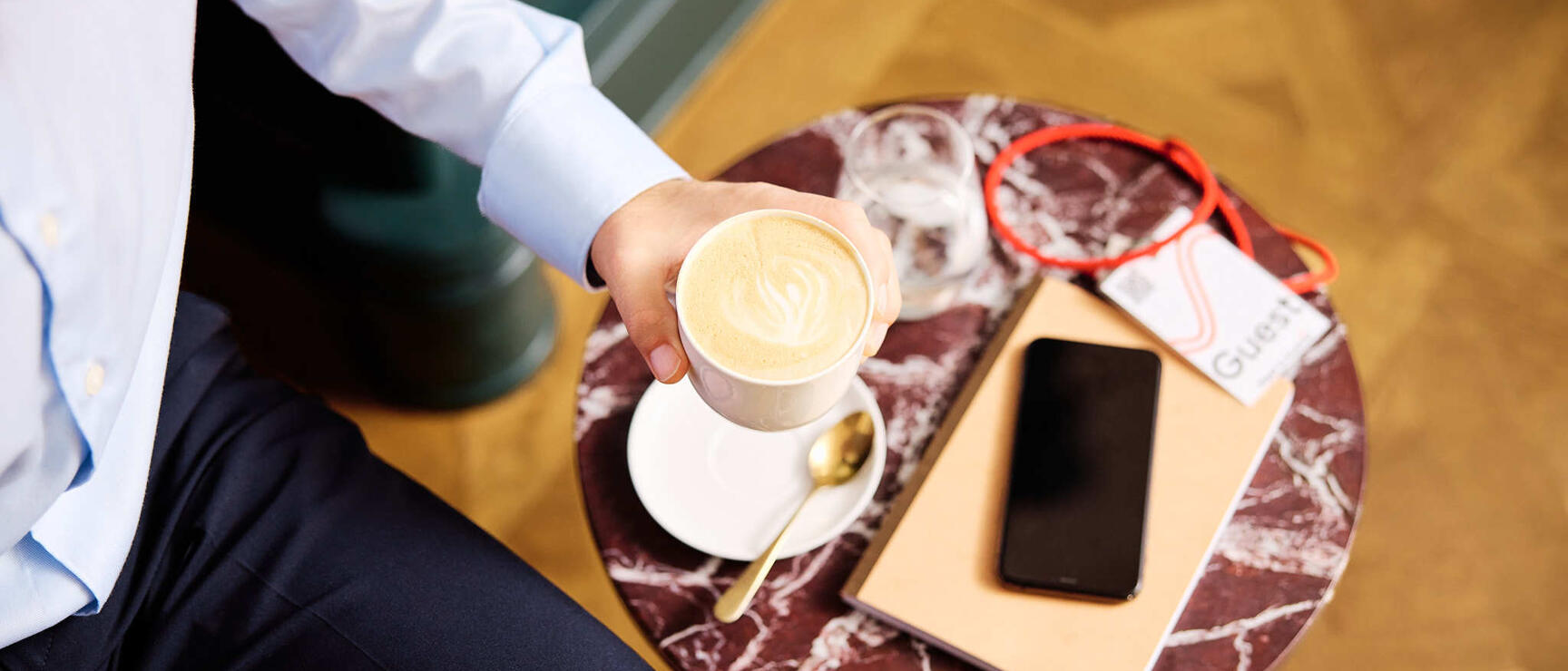Person hält Cappuccino auf rundem Marmortisch mit Smartphone, Notizbuch und roter Brille.