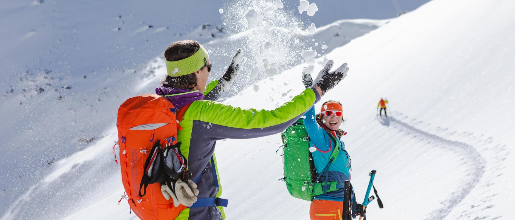 Two ski tourers throwing snow on a snowy mountainside, third person in background.