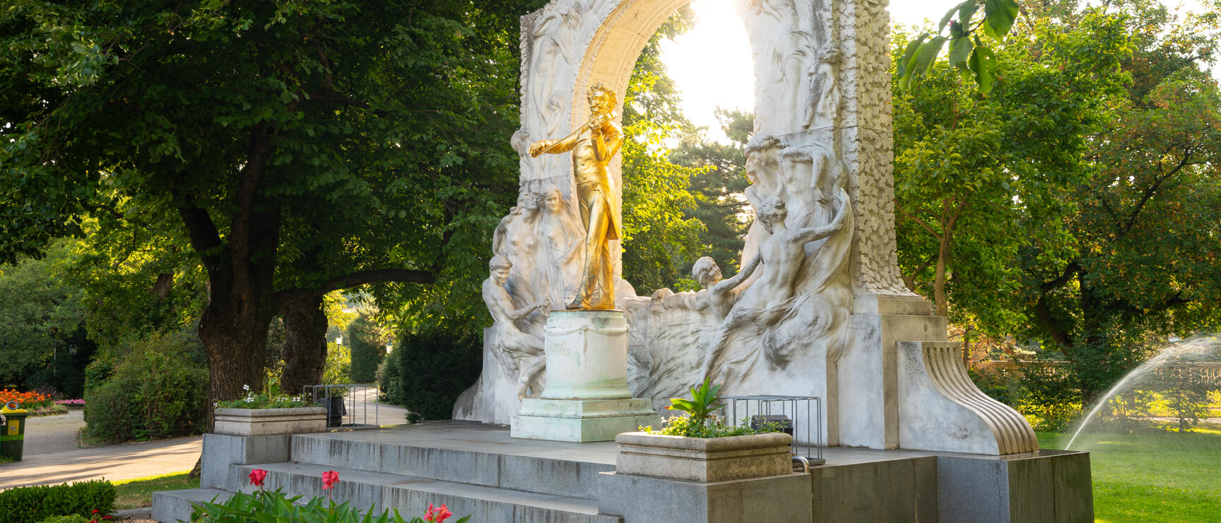 Golden Johann Strauss statue in Vienna's City Park with white marble arch and flowerbeds in foreground.
