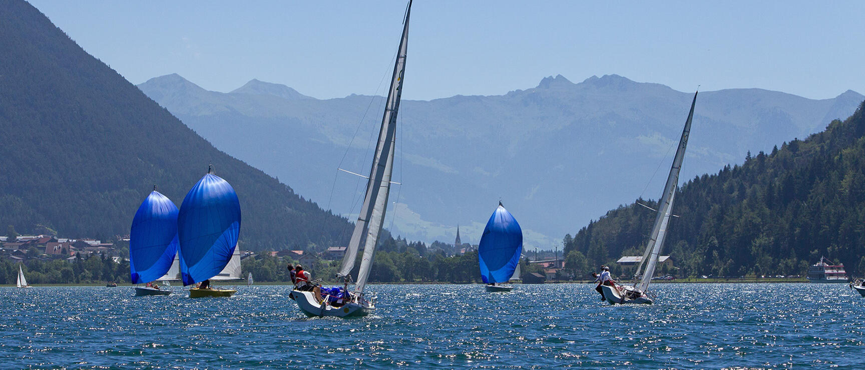 Several sailboats with blue spinnakers on an alpine lake, forested hills and mountain peaks in the background