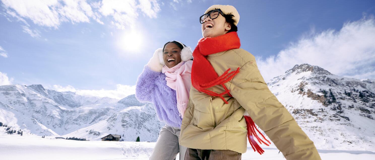 Two smiling people in winter jackets and scarves on snowy mountain landscape in sunshine.