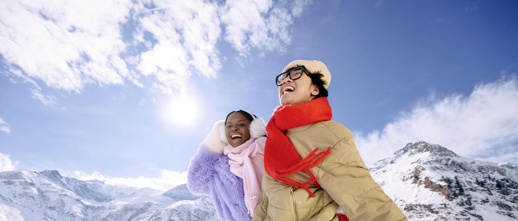 Zwei lachende Personen in Winterjacken und Schals auf schneebedeckter Berglandschaft bei Sonnenschein.
