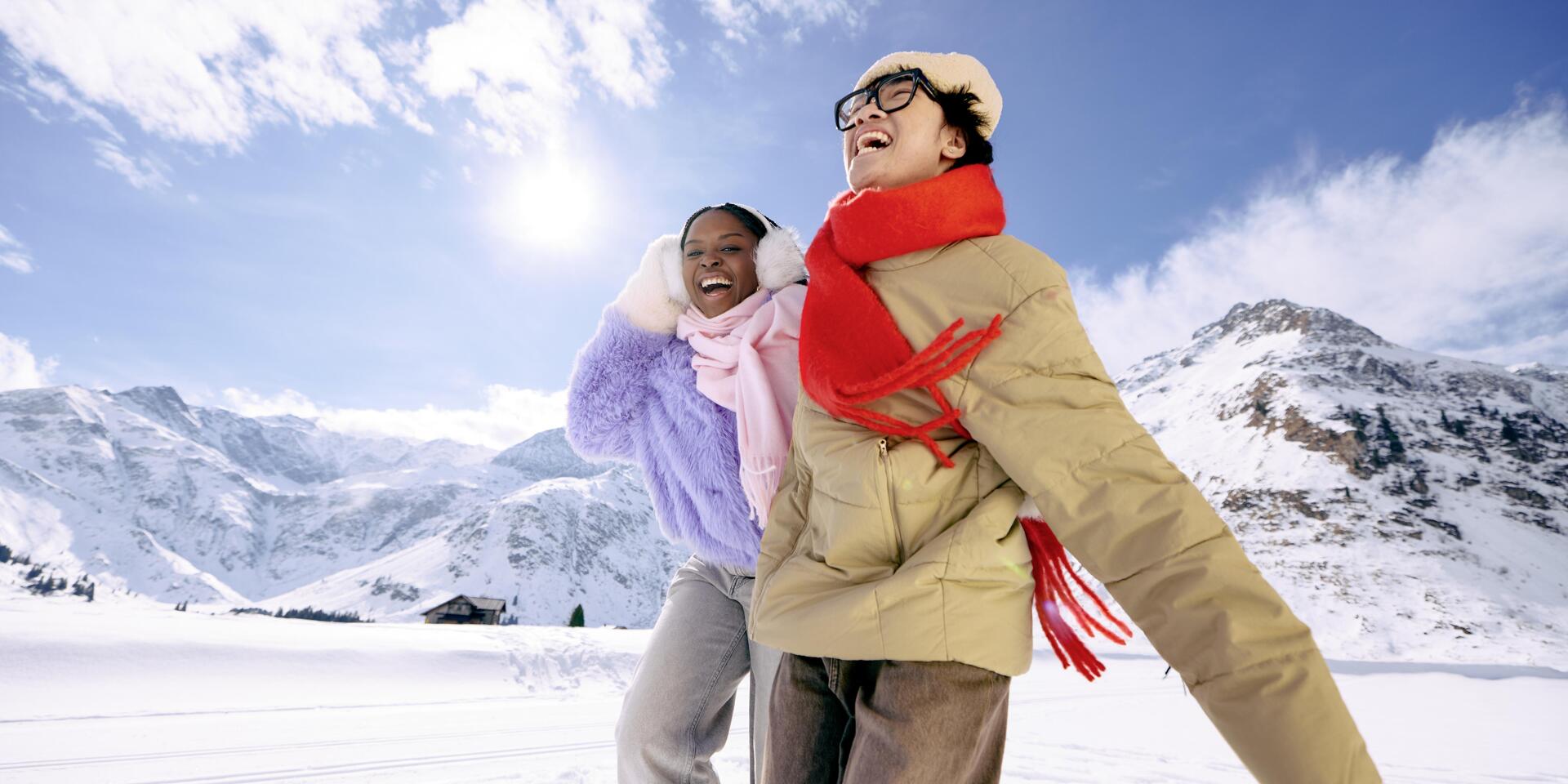 Two smiling people in winter jackets and scarves on snowy mountain landscape in sunshine.