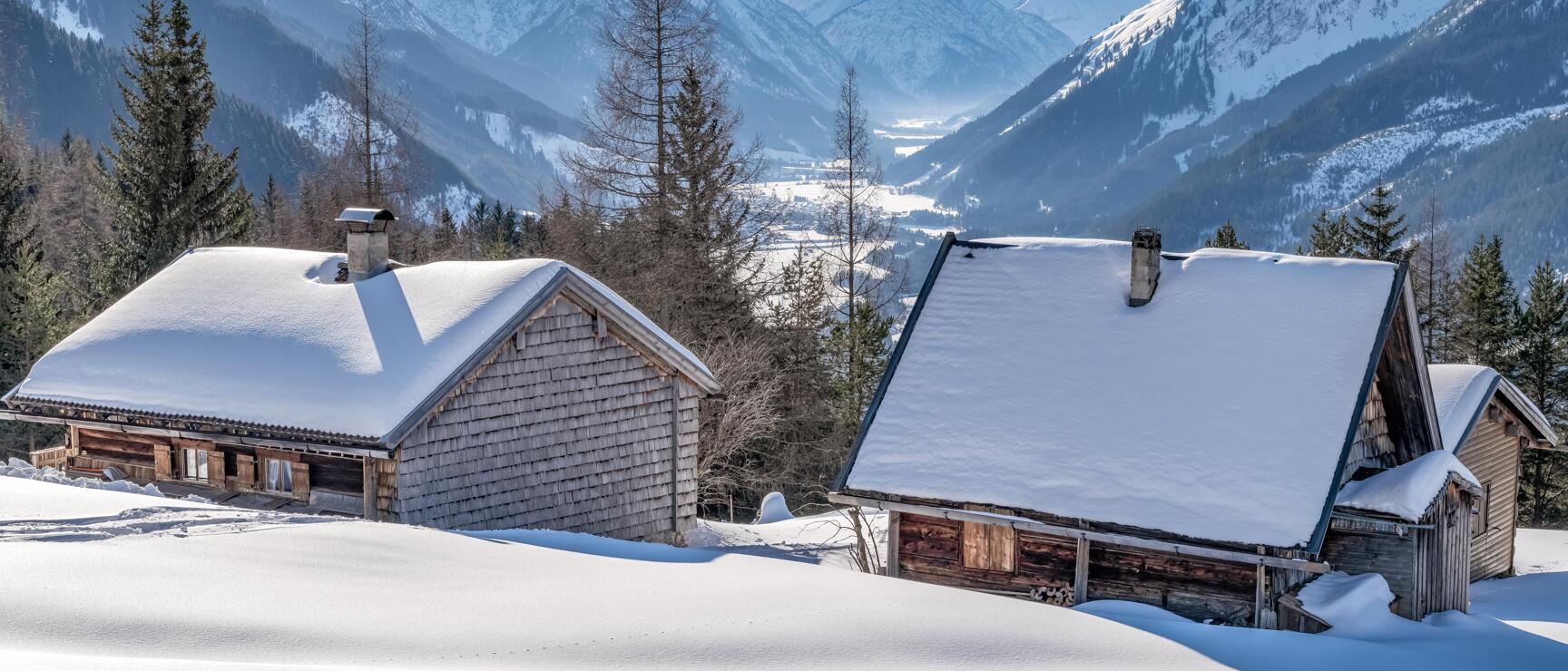 Two snow-covered mountain huts in the Lech Valley, surrounded by spruce trees, with a view of the valley and mountain peaks in the background.