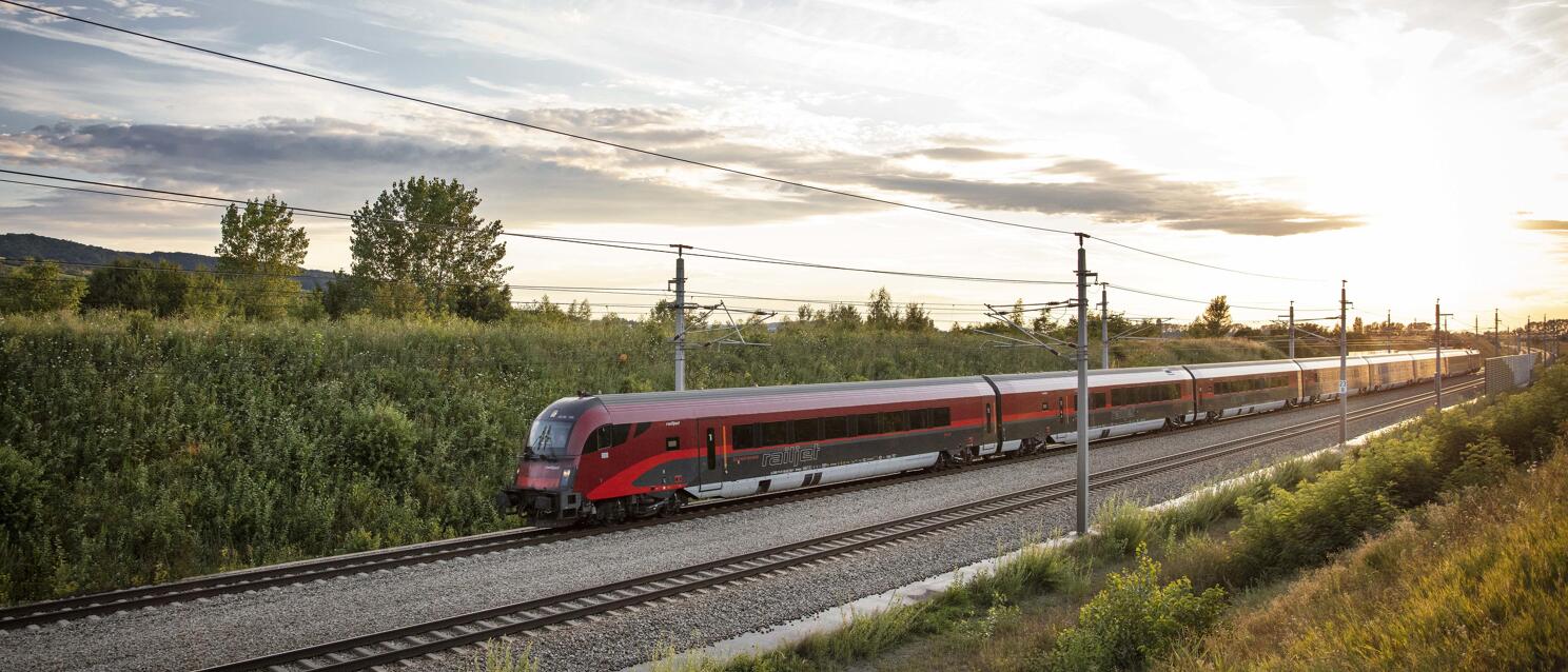 Red Railjet train on double-track line in flat landscape with evening sun in background.