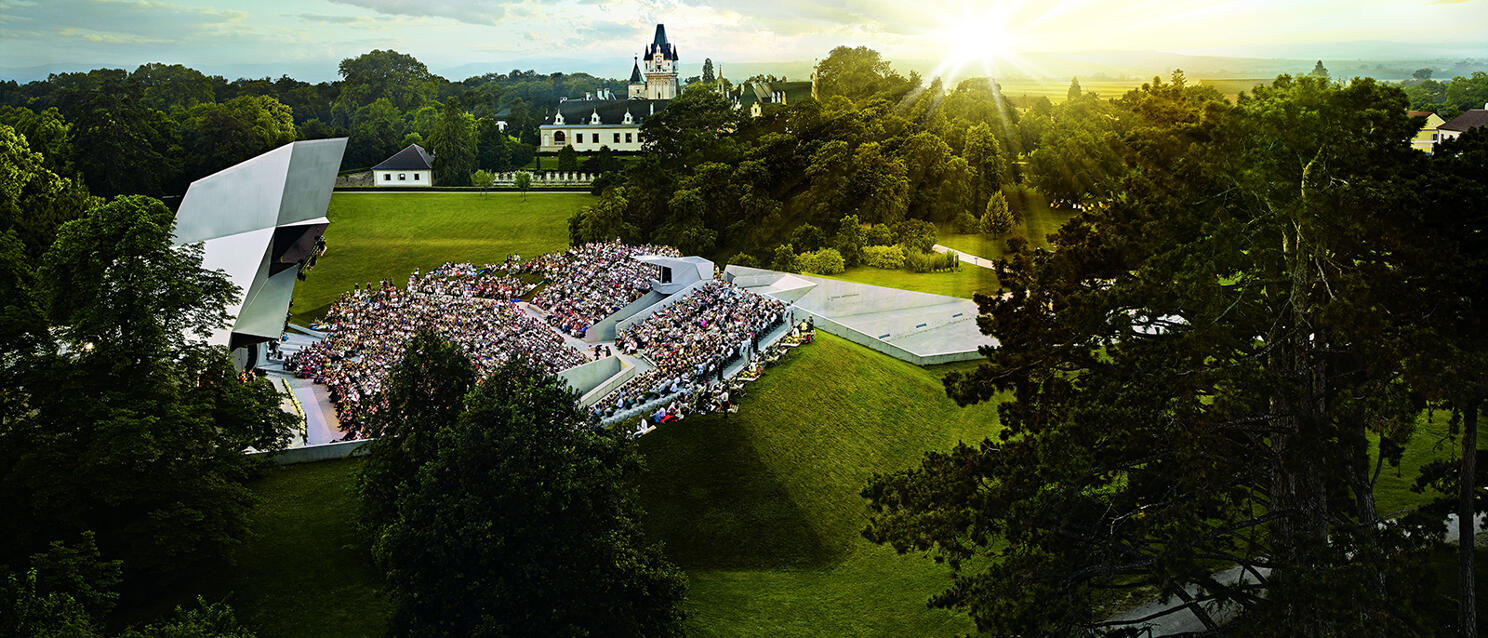 Open-air stage with audience in Schloss Grafenegg park, sunset in background.