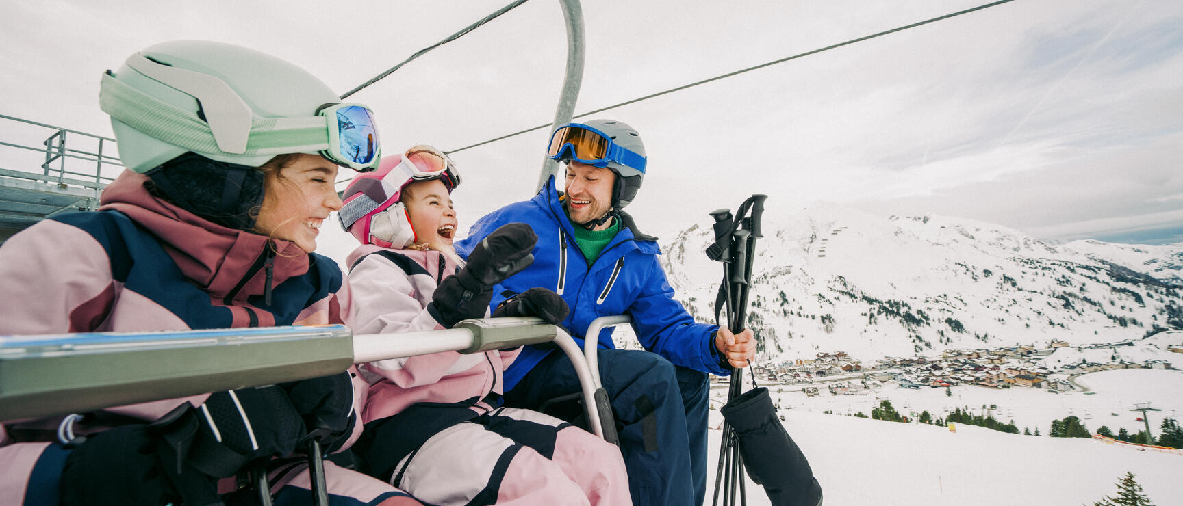 Zwei Erwachsene und ein Kind in Skiausrüstung auf einem Sessellift, schneebedeckte Berge und Ortschaft im Hintergrund.