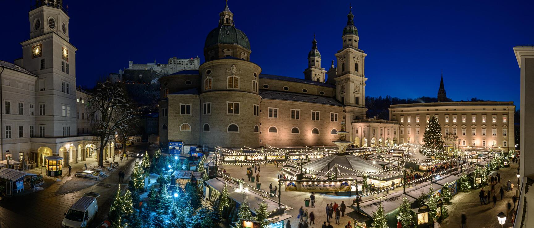 Christmas market at Salzburg Cathedral Square at night with illuminated stalls, Christmas trees, cathedral and fortress in the background.