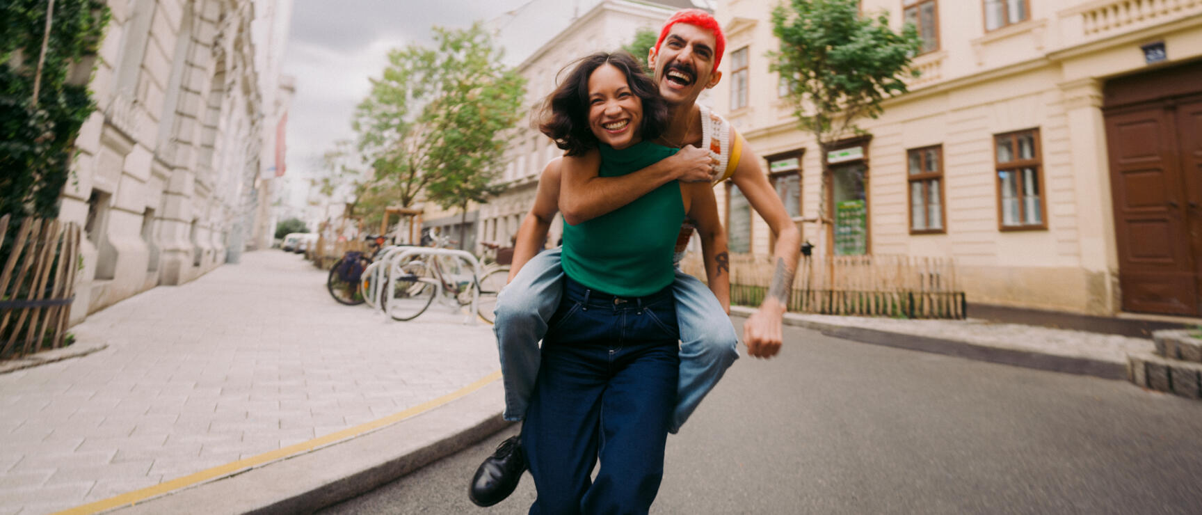 Red-haired man carrying laughing woman piggyback on a Vienna city street.