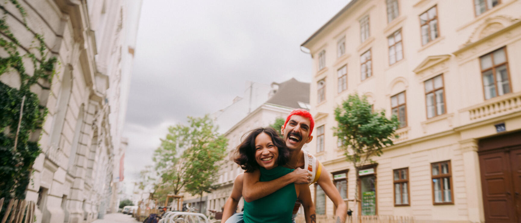 Red-haired man carrying laughing woman piggyback on a Vienna city street.