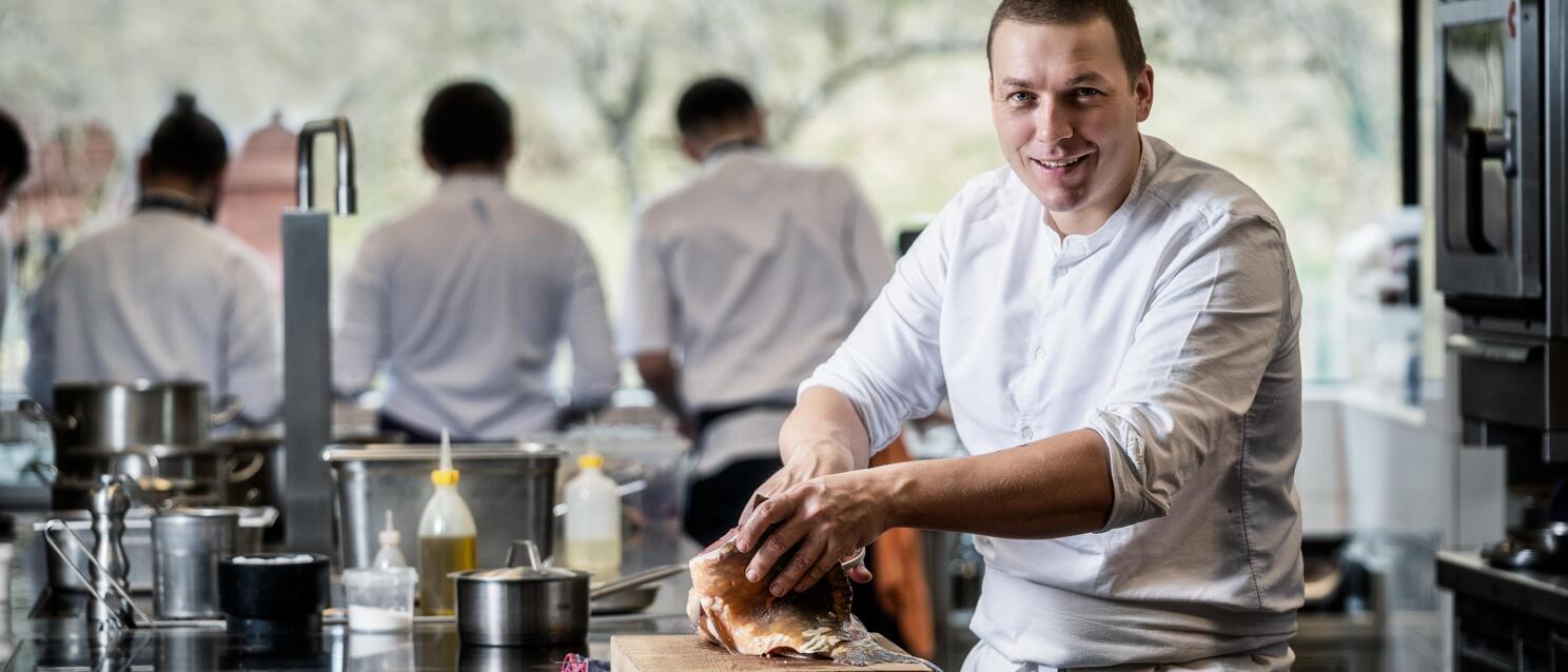 Chef in white jacket working with meat on wooden board, other chefs visible in professional kitchen background.
