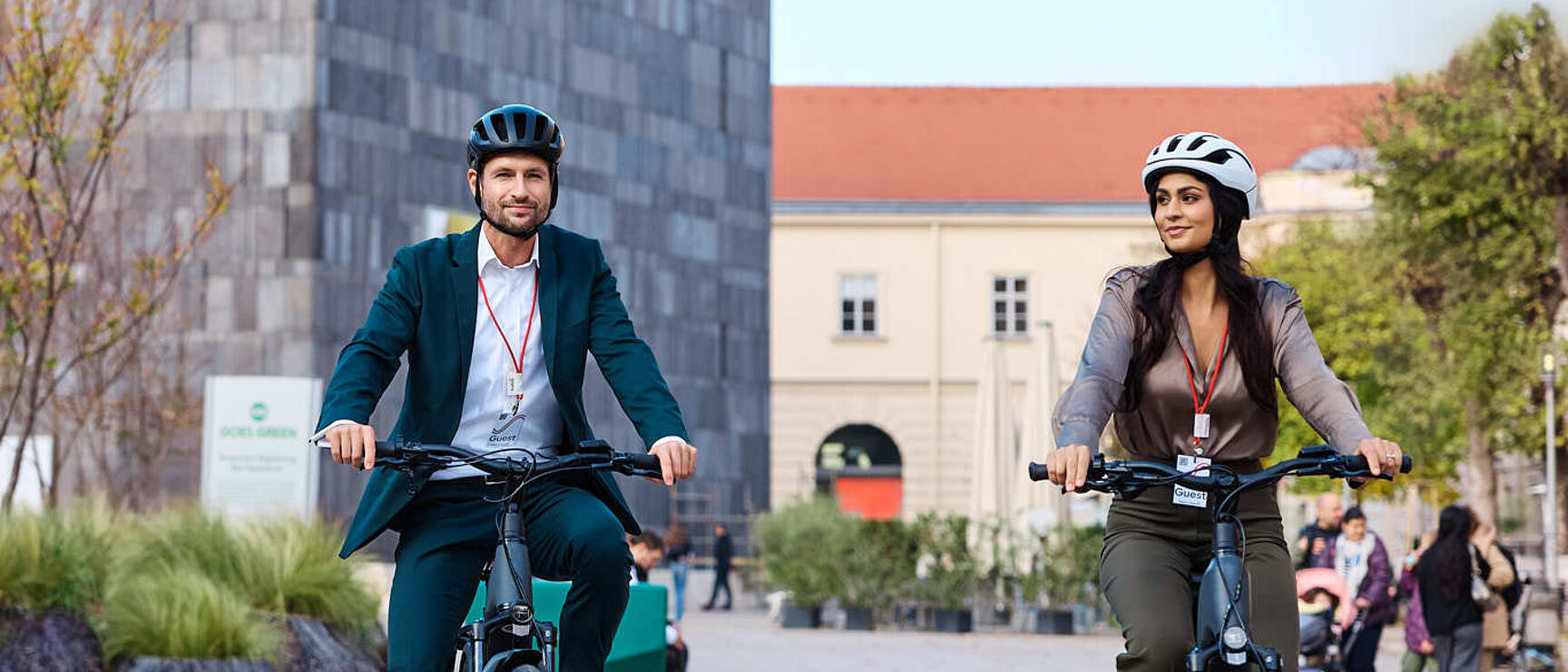 Zwei Personen mit Helmen auf E-Bikes auf einem Stadtplatz, modernes Gebäude im Hintergrund.