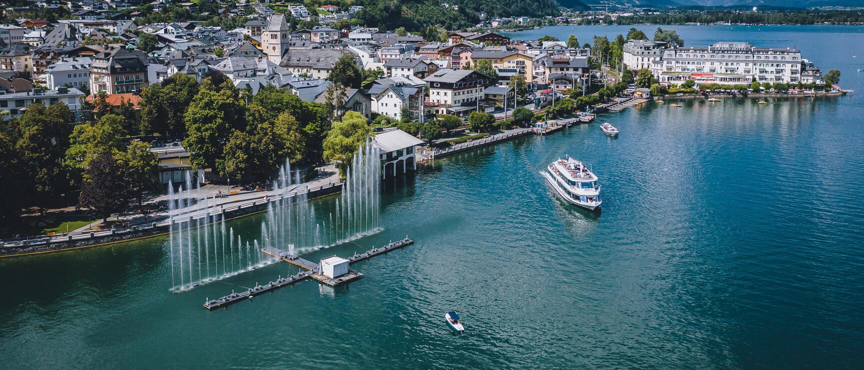 Aerial view of Zell am See with fountain in the lake, excursion boat and mountain panorama in the background.