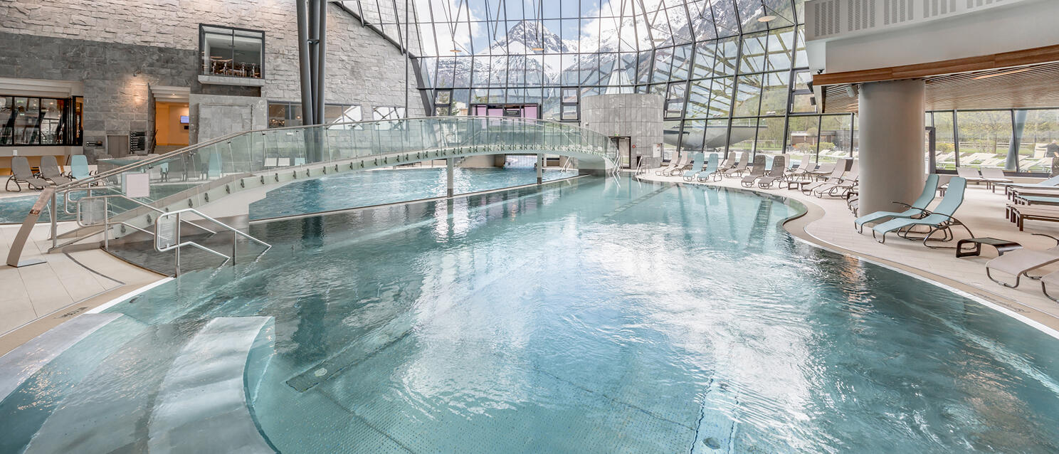 Large indoor swimming pool with glass front, sun loungers and view of snow-capped mountain peaks.