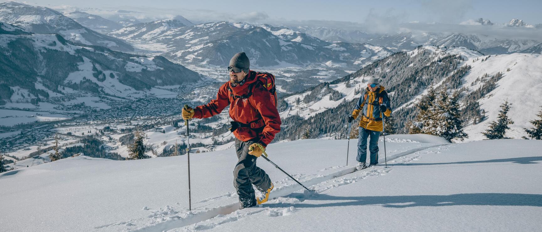 Two ski tourers ascending, snowy mountain landscape with valley and village in the background.