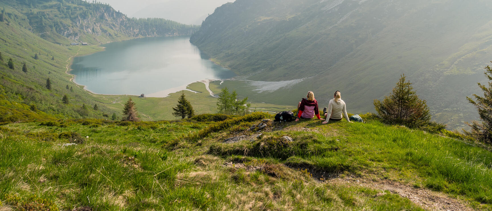 Two women sitting on a mountain meadow overlooking an alpine lake in the valley, mountains in the background.