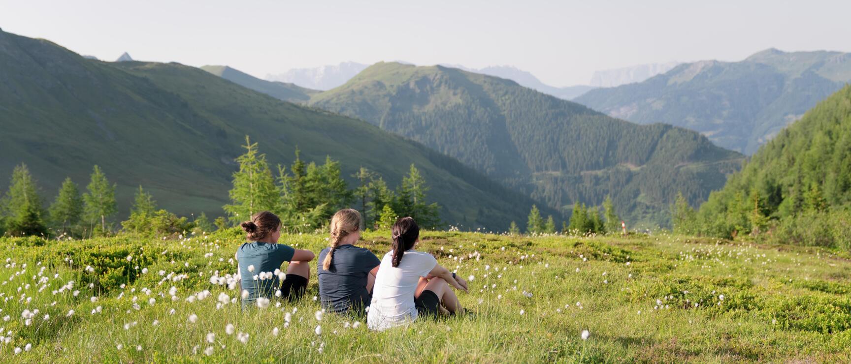Three women sitting on an alpine meadow with cotton grass, view of green mountain slopes in the background.