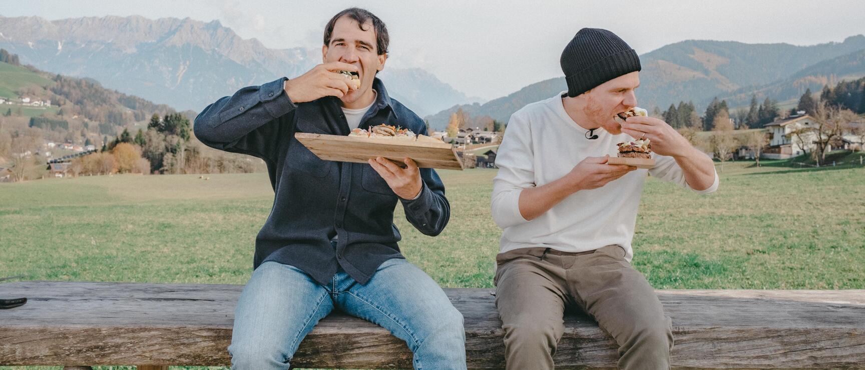 Two men eating burgers on wooden bench, green meadow and mountain panorama in background.