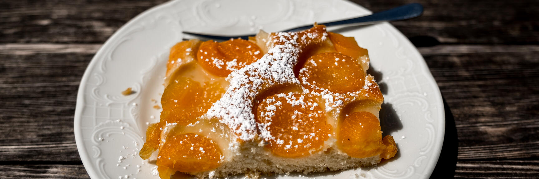 Slice of apricot cake with icing sugar on white plate, fork, rustic wooden table.