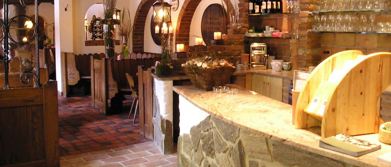 Rustic inn interior with stone counter, wooden beams, brick arches, wine bottles and glasses on shelves.