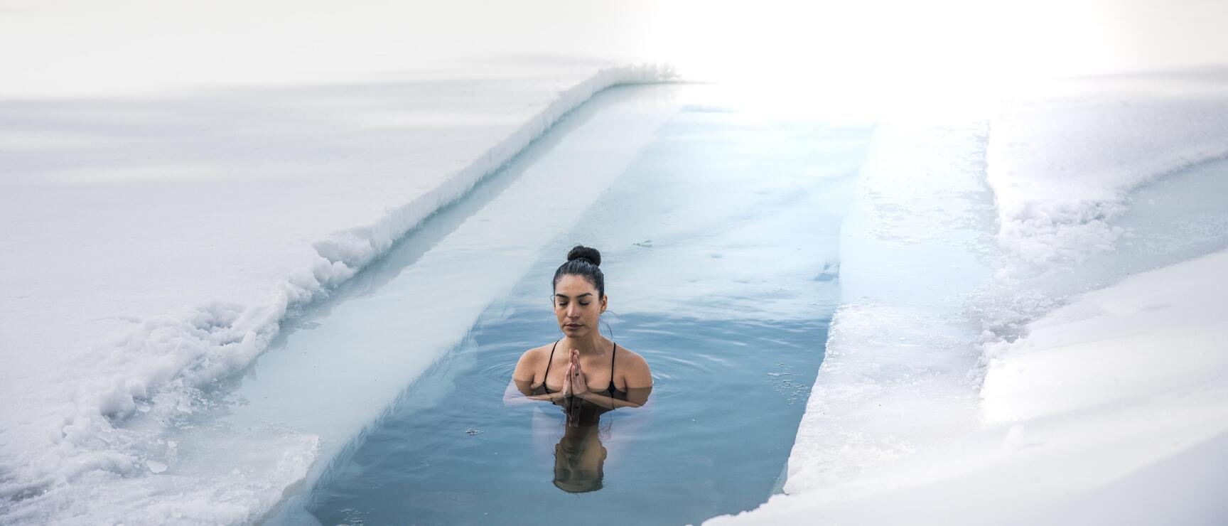 Frau badet in einem Eisloch in verschneiter Landschaft, schneebedeckte Berge im Hintergrund.