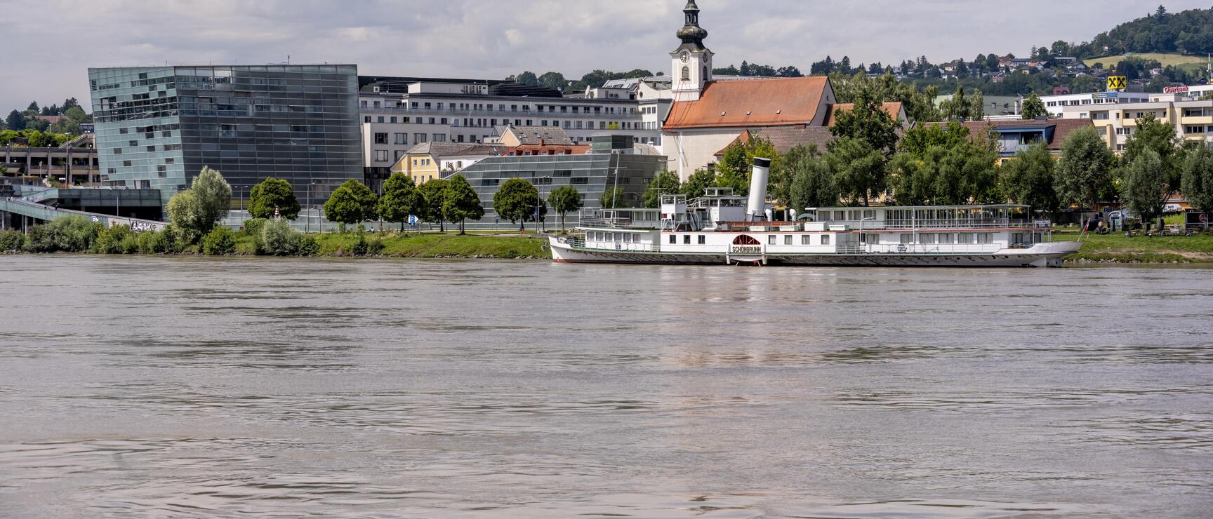View across the Danube to Linz with church tower, modern glass building and moored steamship.