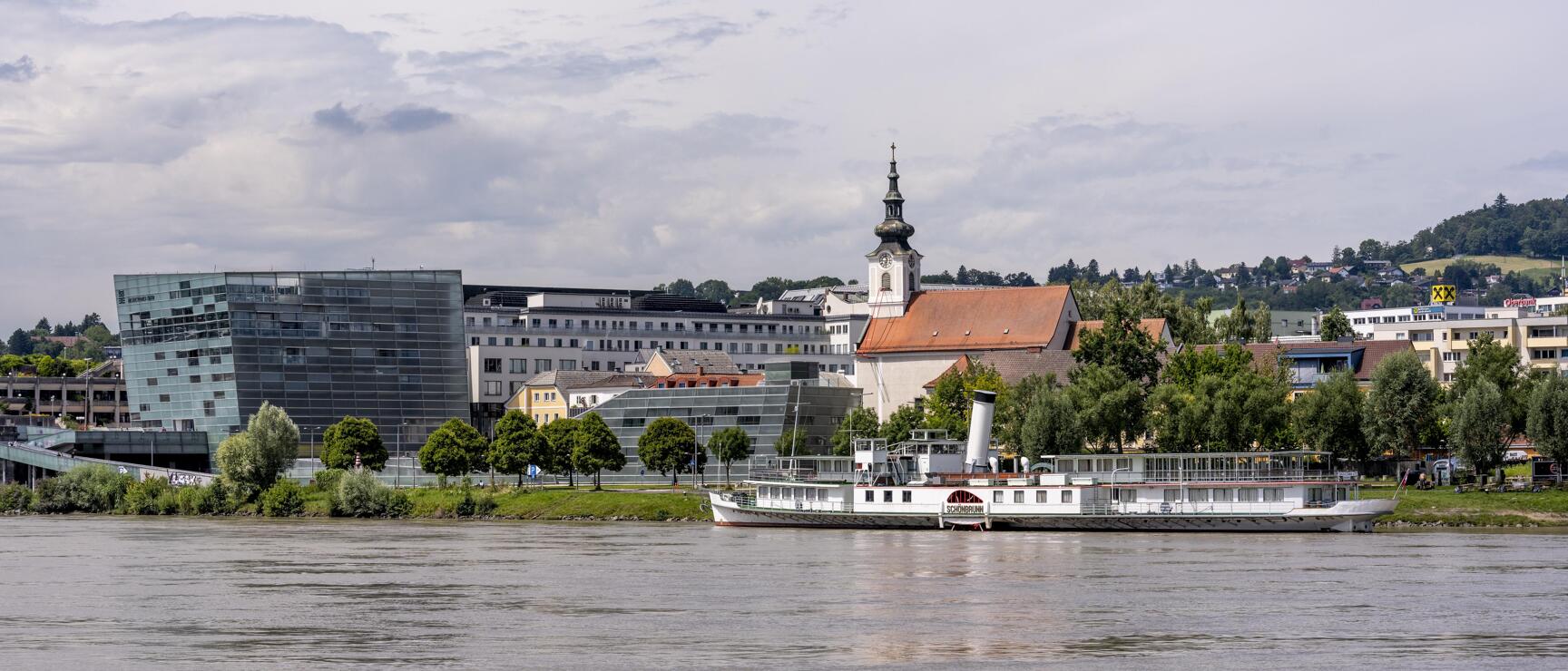 View across the Danube to Linz with church tower, modern glass building and moored steamship.
