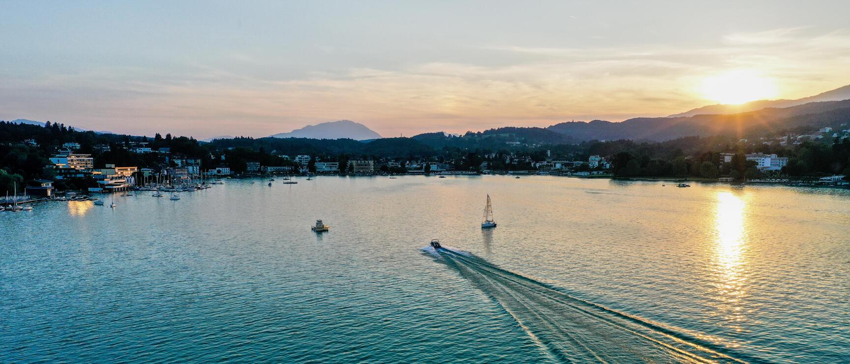 Motor boat with wake and sailing boat on lake at sunset, village and mountains in background.