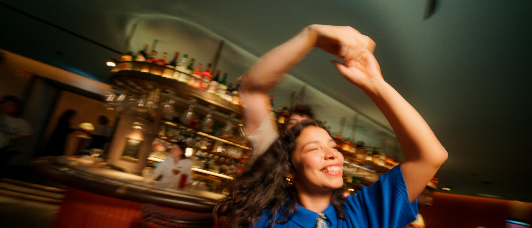 Woman dancing in blue top at bar with bottle rack and bartender in background.
