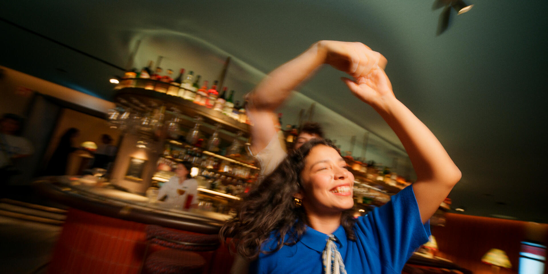 Tanzende Frau im blauen Oberteil in einer Bar mit Flaschen-Regal und Barkeeper im Hintergrund.