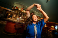 Tanzende Frau im blauen Oberteil in einer Bar mit Flaschen-Regal und Barkeeper im Hintergrund.