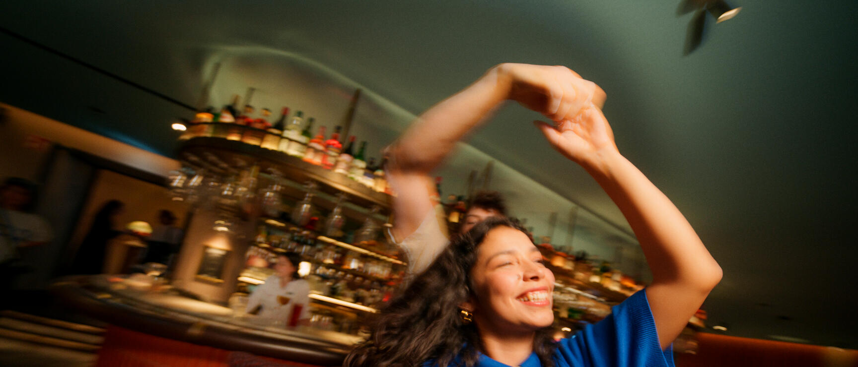 Tanzende Frau im blauen Oberteil in einer Bar mit Flaschen-Regal und Barkeeper im Hintergrund.