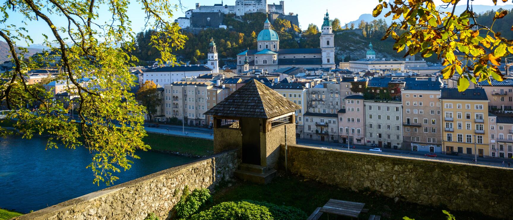 View of Salzburg's old town with cathedral and Hohensalzburg Fortress, autumn trees in foreground.