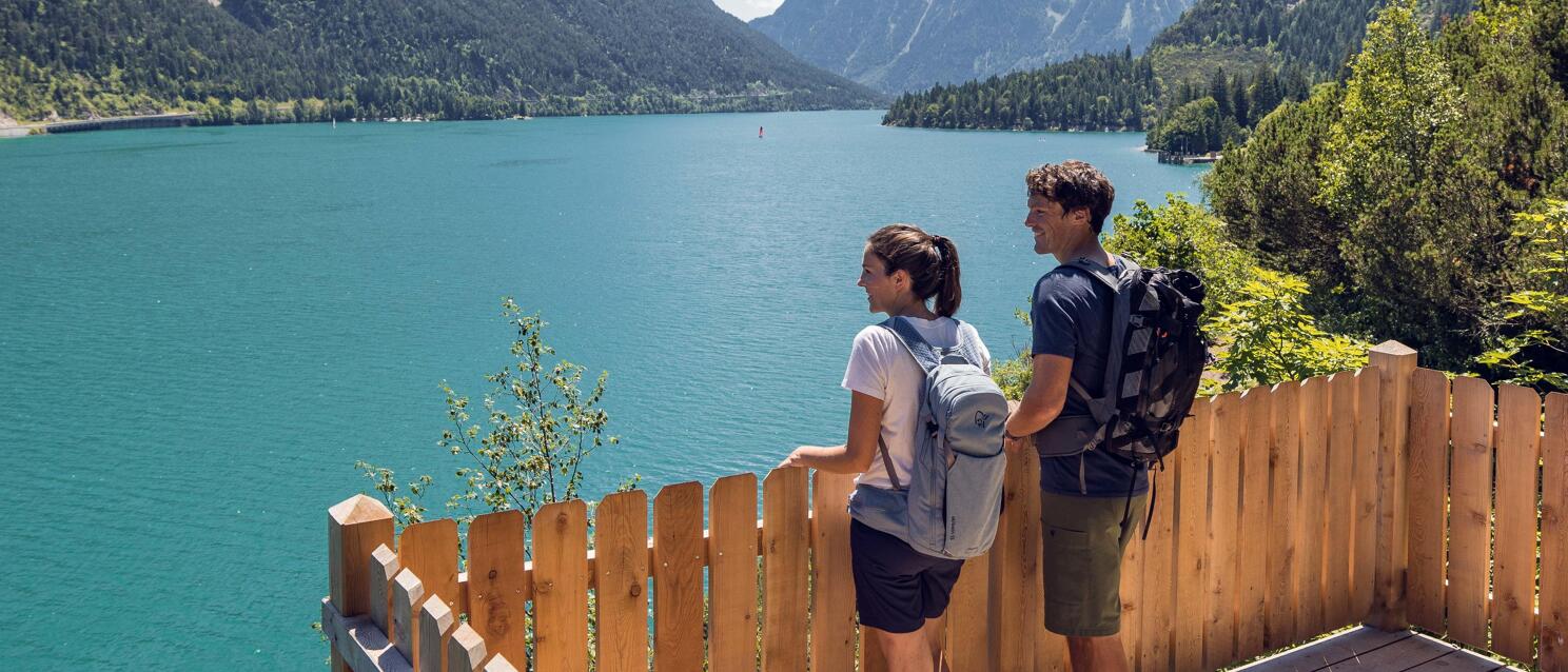 Two hikers with backpacks on a wooden terrace overlooking the turquoise Achensee lake in Tyrol.