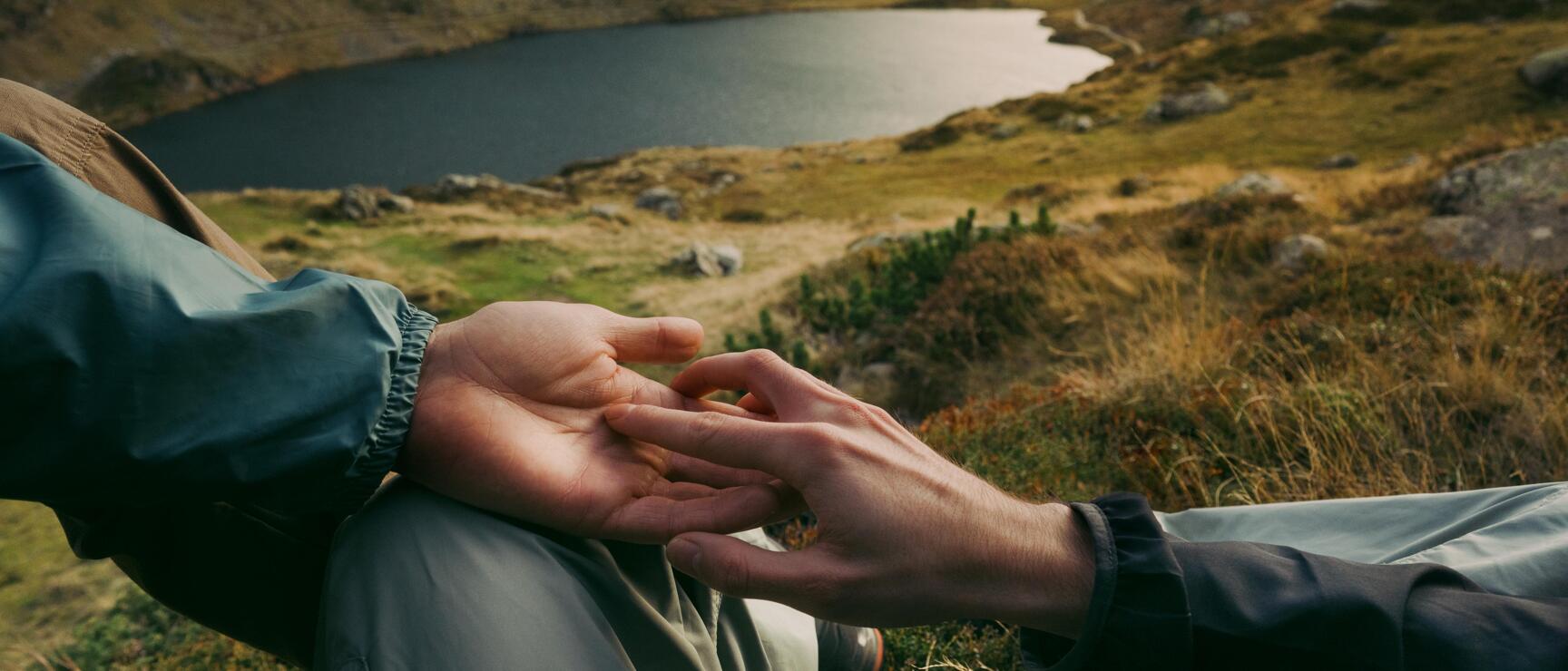 Two hands touching in foreground, alpine mountain lake in background.